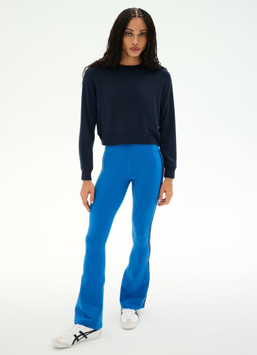 A model wearing a dark navy crewneck sweatshirt, bright blue flared pants, and white sneakers, standing against a plain white studio background.