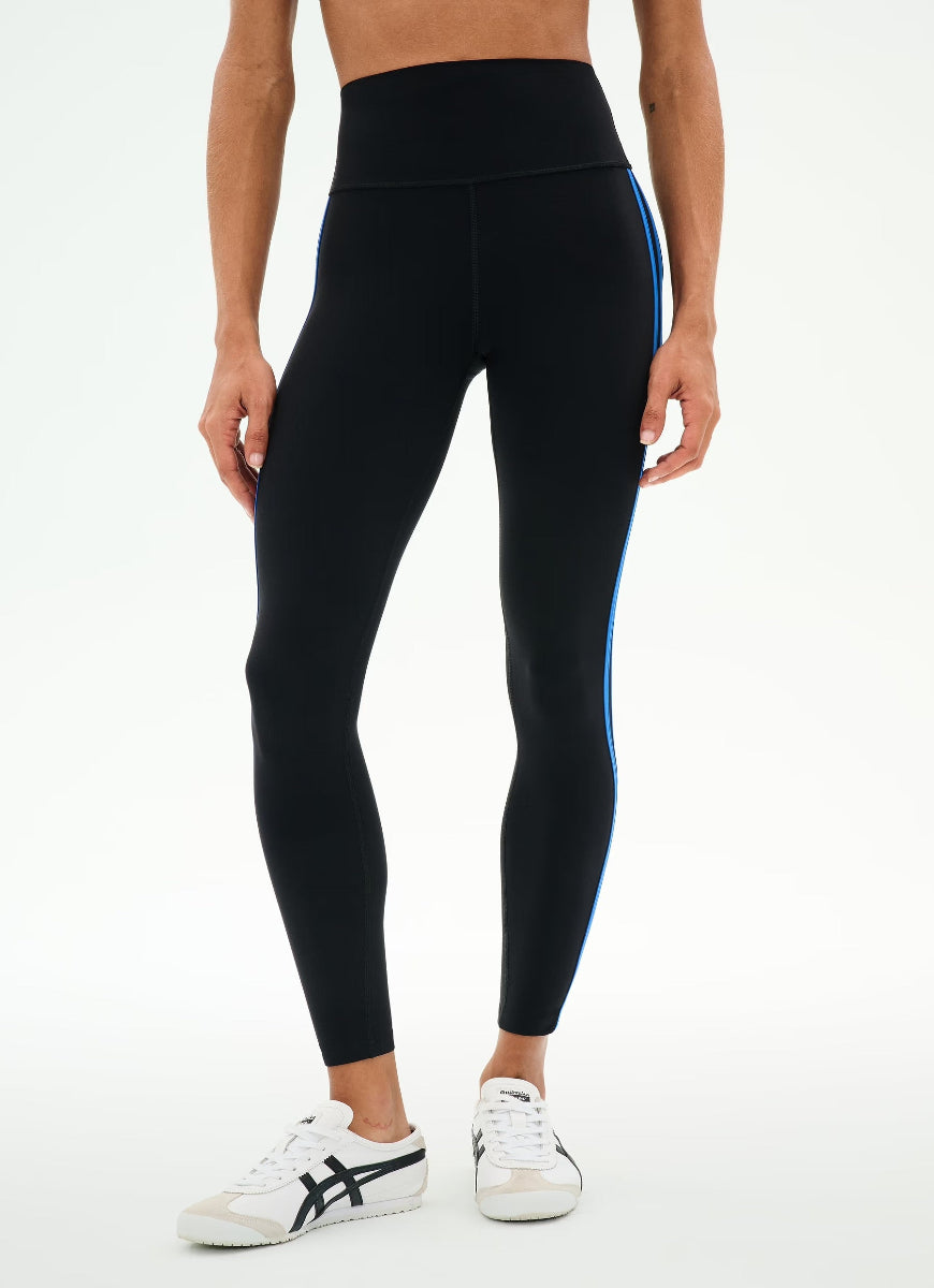 A model wearing black high-waisted leggings with a bright blue side stripe, paired with white sneakers, shown from the waist down against a plain white studio background.