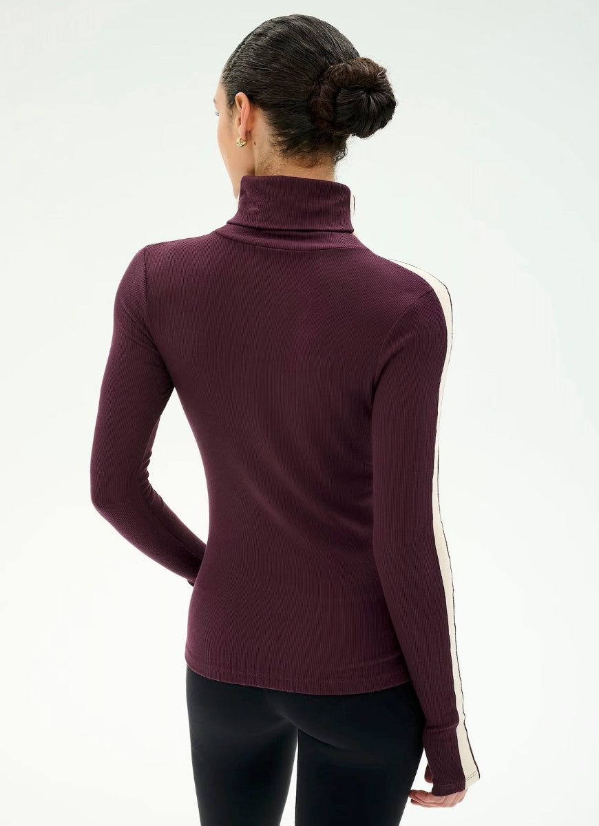 A woman viewed from the back wearing a fitted long-sleeve turtleneck top in deep burgundy with a white stripe running down the sleeve, paired with black leggings, against a plain white studio background.