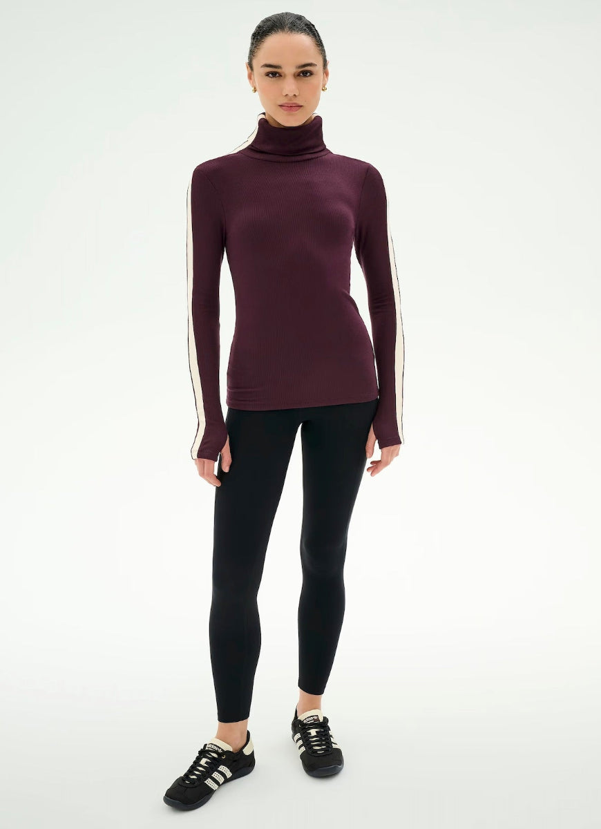 A model wearing a dark burgundy long-sleeve turtleneck top with white stripes down the sleeves, paired with black leggings and black sneakers, against a plain white studio background.