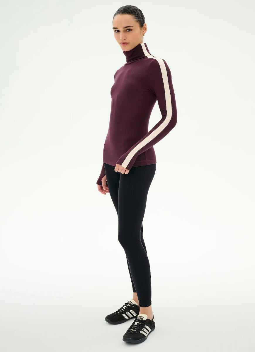 A woman modeling a long-sleeve turtleneck top in deep burgundy with cream stripes down the sleeves, paired with black leggings and black sneakers, against a plain white studio background.
