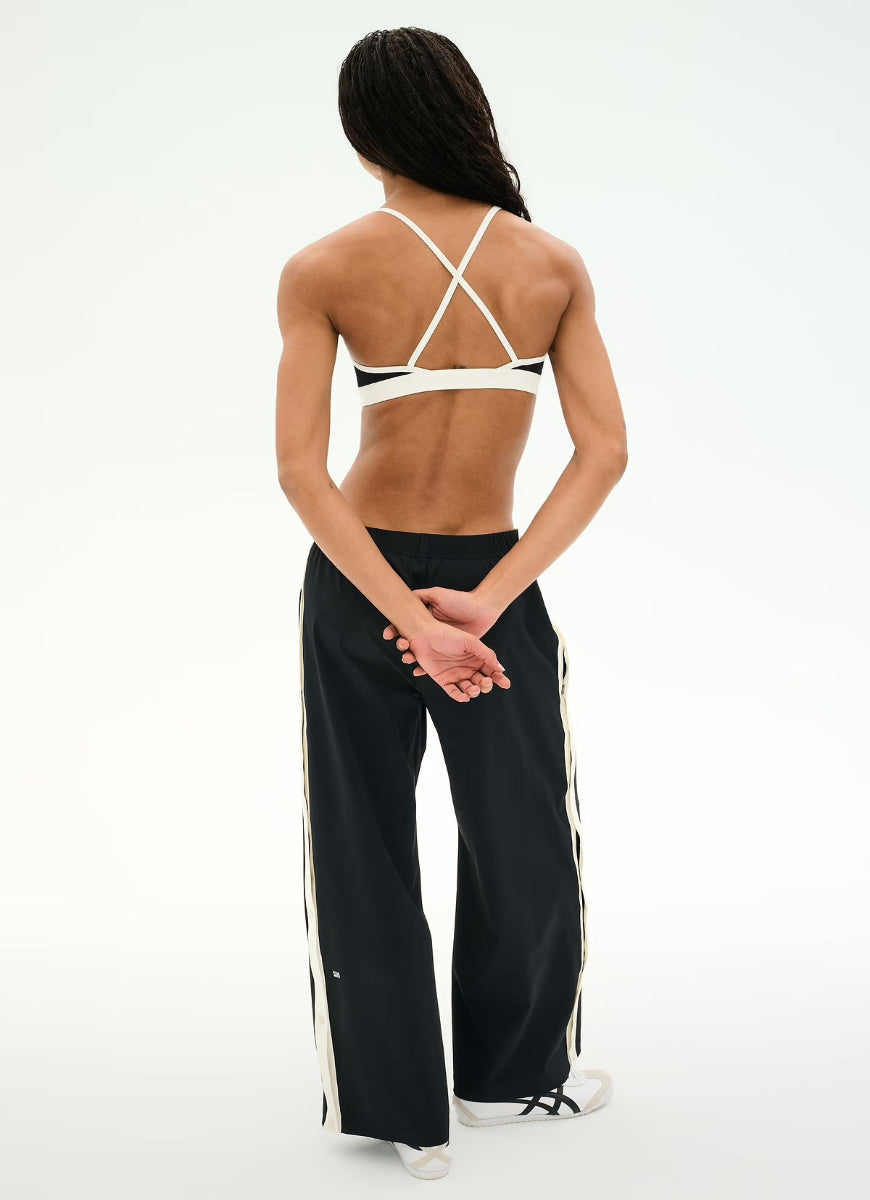 A woman shown from the back wearing black wide-leg track pants with white side stripes, a white strappy sports bra, and white sneakers against a clean white studio background.