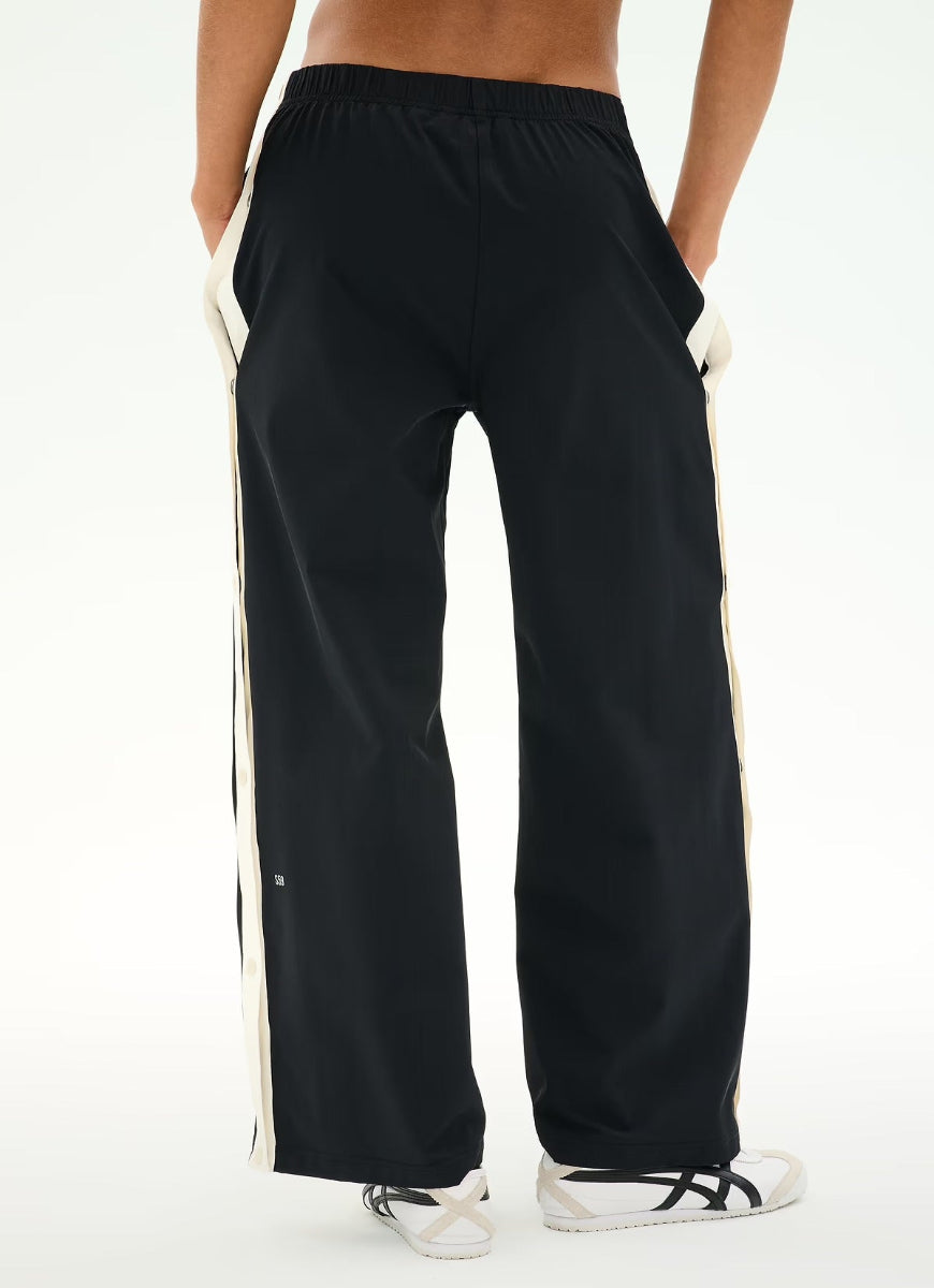 Black wide-leg track pants with cream side stripes, shown on a model from the back against a white studio background.
