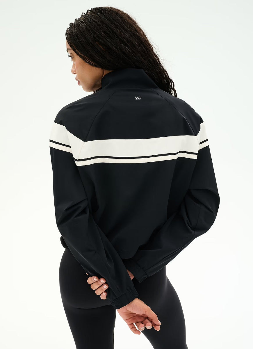 A woman shown from the back wearing a black athletic jacket with two horizontal white stripes across the upper back and shoulders, paired with black leggings, against a plain white studio background.