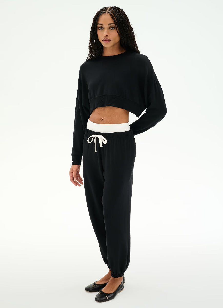 A woman modeling a black two-piece loungewear set consisting of a cropped long-sleeve top and matching jogger pants with a white drawstring waistband, photographed against a plain white studio background.