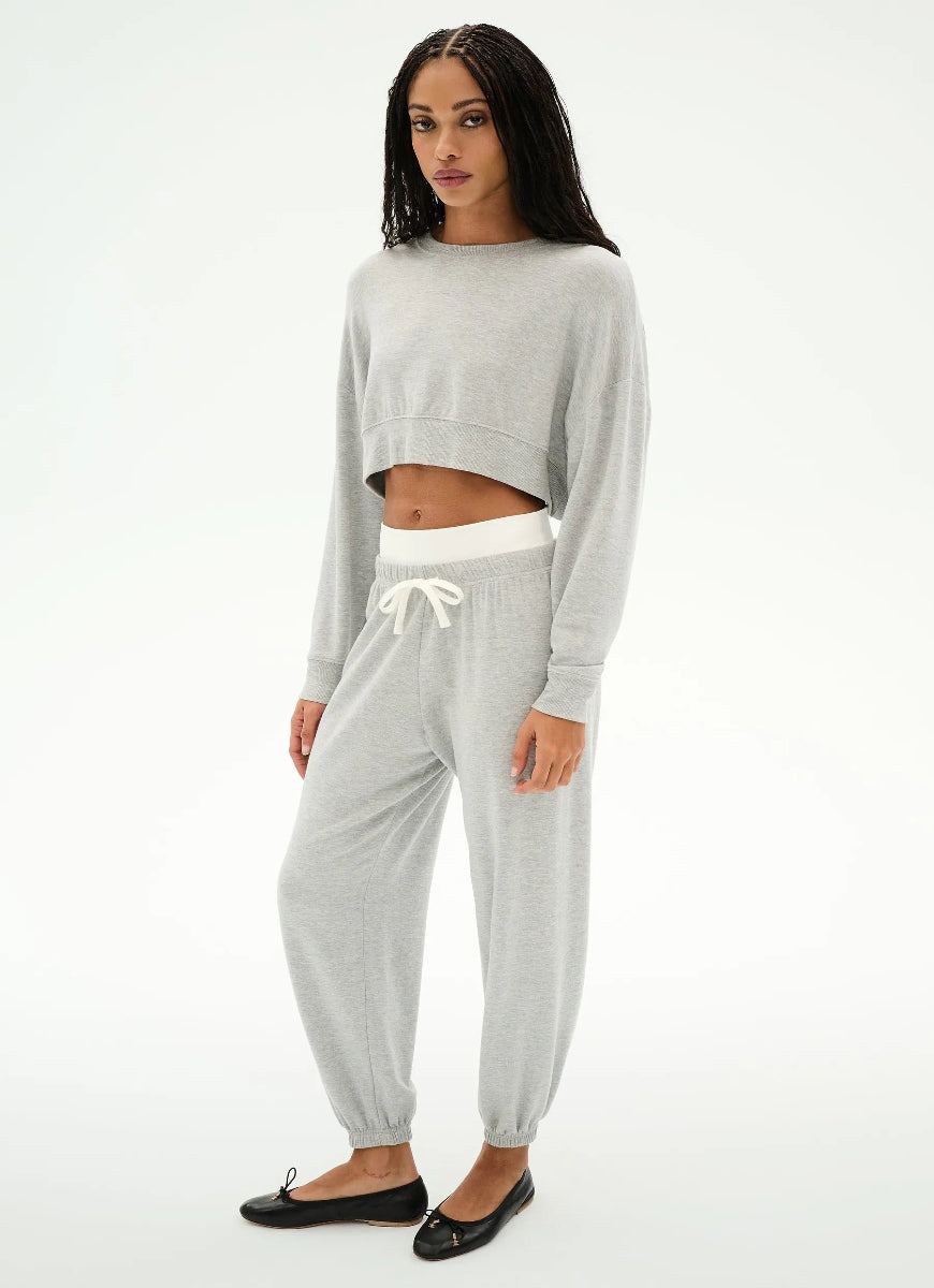 A woman modeling a light gray two-piece loungewear set consisting of a cropped long-sleeve sweatshirt and matching jogger pants with a white drawstring waistband, paired with black flats, against a plain white studio background.
