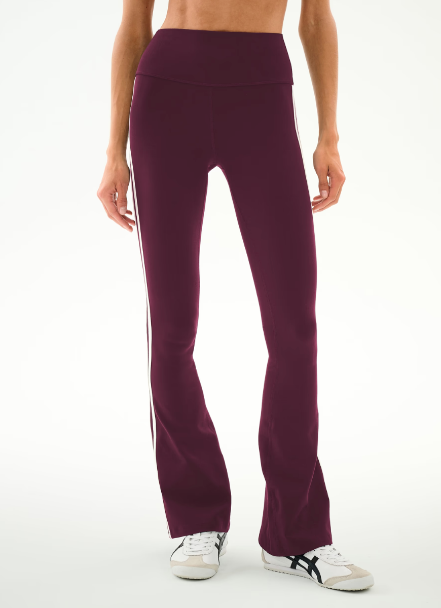 A model from the waist down wearing high-waisted, wide-leg athletic pants in a deep plum purple color, featuring white side stripes, paired with white sneakers.