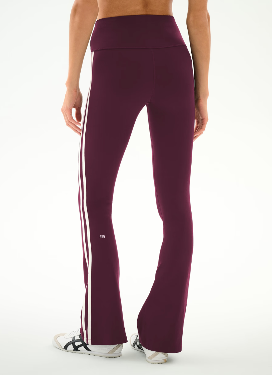 A person shown from the back wearing high-waisted flared leggings in deep burgundy with two white side stripes and white sneakers, photographed against a clean white studio background.