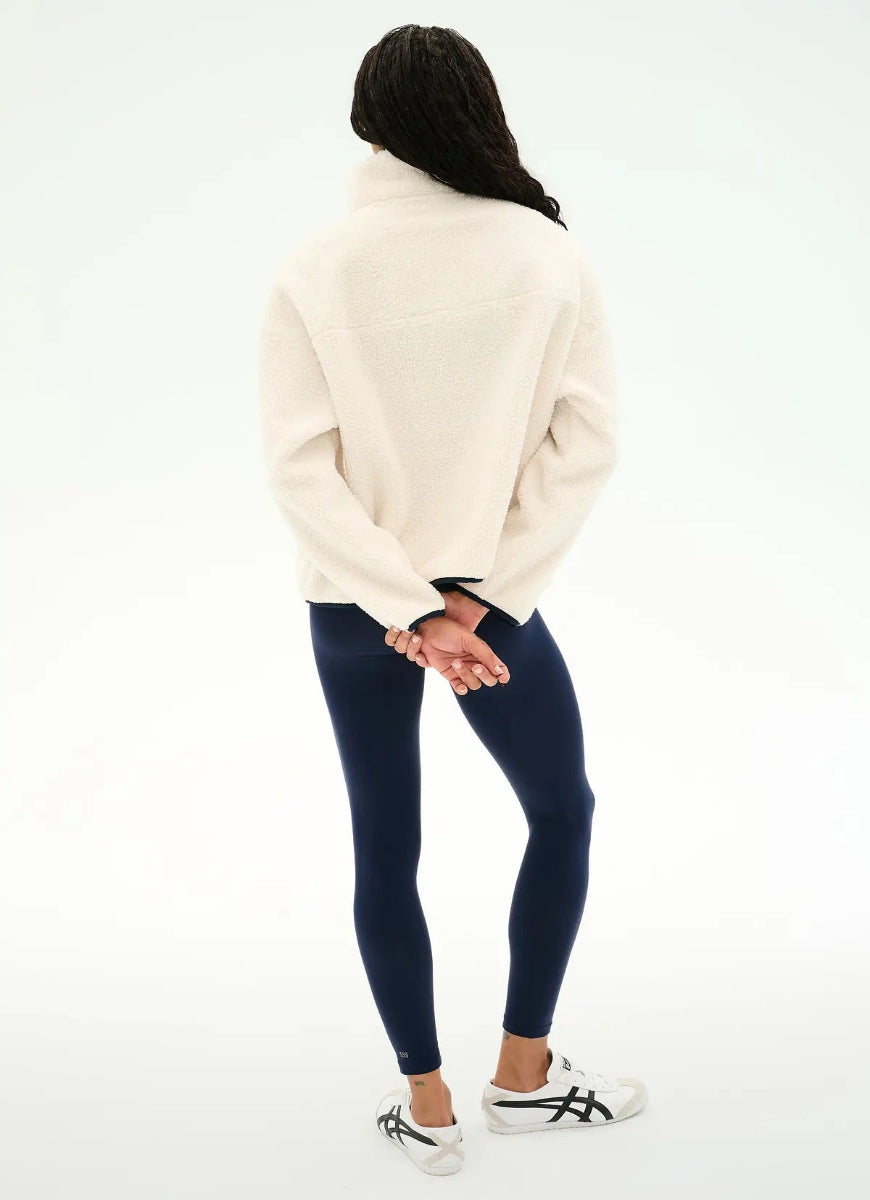 A woman shown from the back wearing a cream-colored fuzzy mock-neck sweater, dark navy leggings, and white sneakers with black stripes, set against a plain white studio background.