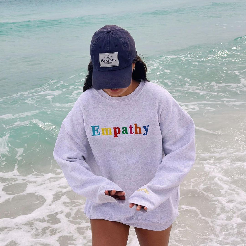 Model on the Beach Wearing The Mayfair Group Empathy Always Crewneck in Grey