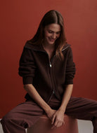 A woman wearing a dark brown ribbed zip-up hoodie and matching sweatpants, sitting on a stool against a solid terracotta background.