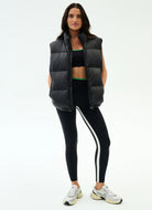 Woman wearing a black puffer vest, black sports bra, and black leggings with white stripes on a white background