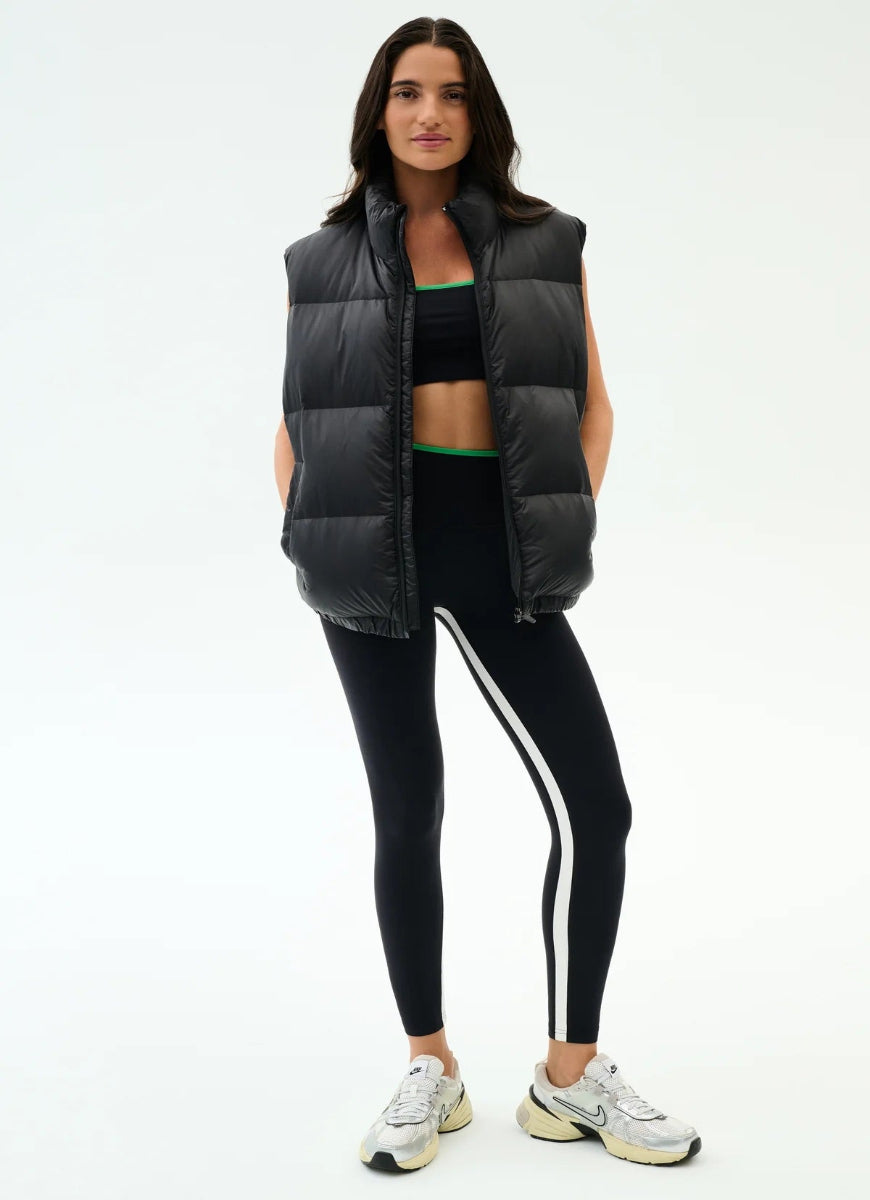 Woman wearing a black puffer vest, black sports bra, and black leggings with white stripes on a white background