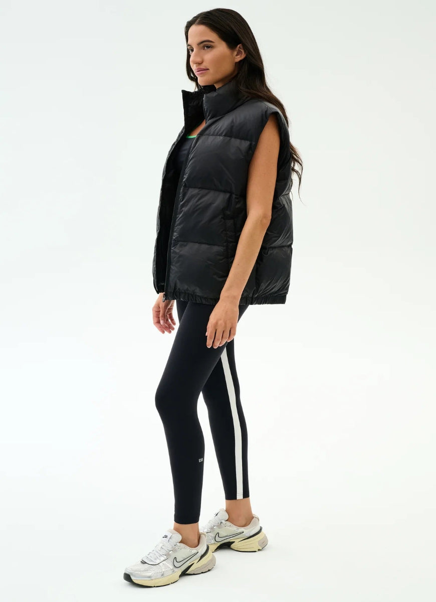 Woman wearing a black puffer vest, black leggings with white stripes, and white sneakers on a white background