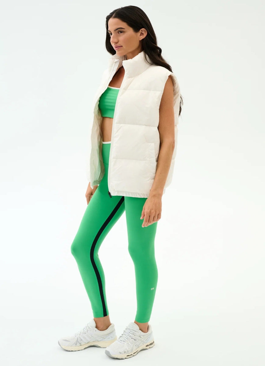 Woman wearing a white puffer vest, green sports bra, and green leggings with black stripes on a white background