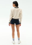 Person wearing a cream sweater with black and white striped trim, navy shorts, and white shoes on a white background
