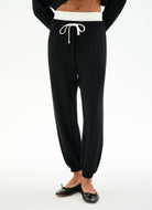 Black jogger pants with a white drawstring and a white waistband, shown on a model wearing black flats against a white studio background.