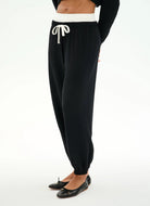 Black jogger pants with a white elastic waistband and white drawstring, shown on a model from the waist down wearing black flats against a white studio background.