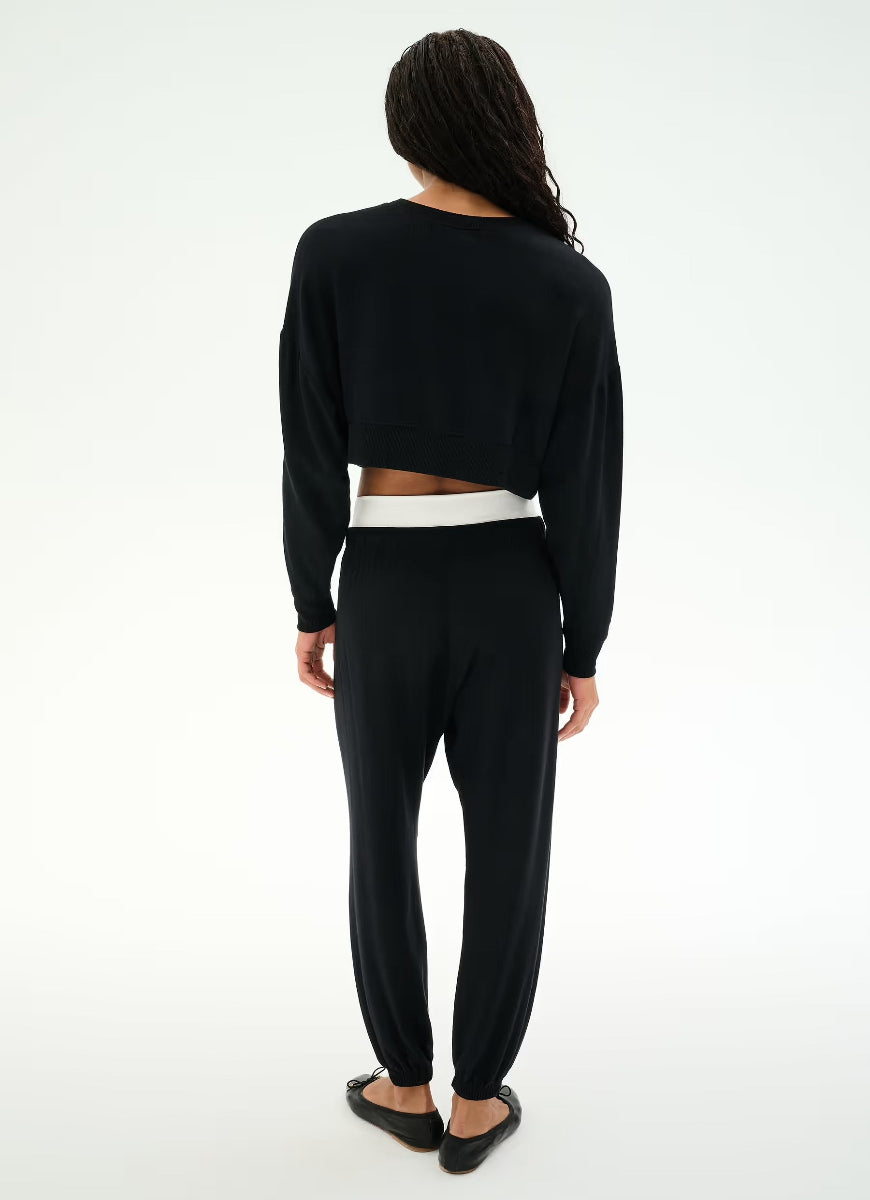 A woman shown from the back wearing a black long-sleeve cropped top with a white underlayer peeking out at the waist, paired with black jogger pants and black flats, against a plain white studio background.