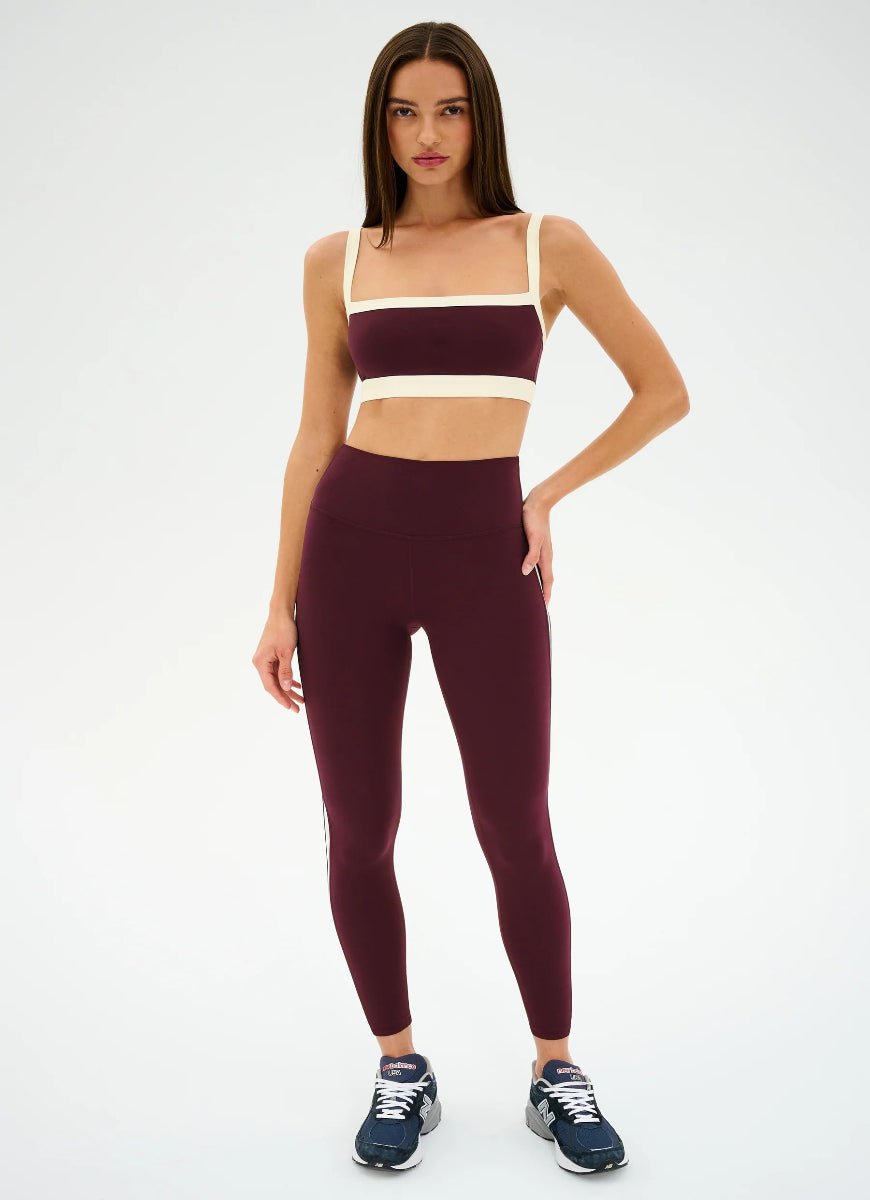 Woman wearing a burgundy sports bra and leggings with a white stripe on a light gray background