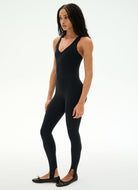 A woman modeling a black sleeveless bodysuit with integrated leggings and black ballet flats, photographed against a clean white studio background.