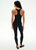 A woman shown from the back wearing a black sleeveless bodysuit with a racerback-style open back and matching black leggings, photographed against a clean white studio background.
