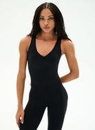 A woman modeling a black sleeveless bodysuit or jumpsuit with a deep V-neck and full-length legs, photographed against a clean white studio background.