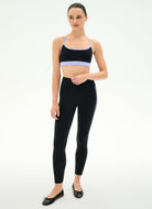 A woman modeling a matching black activewear set consisting of a sports bra with light purple trim and high-waisted leggings with white side stripes, paired with black flats, against a plain white studio background.
