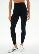 A model wearing black high-waisted leggings with a bright blue side stripe, paired with white sneakers, shown from the waist down against a plain white studio background.