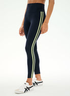 A model wearing navy blue high-waisted athletic leggings with two vertical lime green stripes down the sides, paired with white sneakers, against a plain white background.