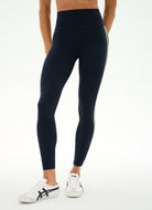 A model from the waist down wearing high-waisted navy blue leggings with a thin yellow-green side stripe, paired with white sneakers, against a plain white background.