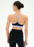 A woman shown from the back wearing a black sports bra with light purple straps and waistband, paired with black leggings featuring white side stripes, against a plain white studio background.