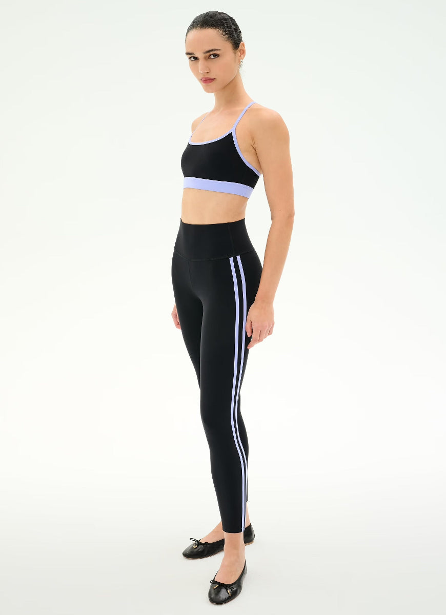 A woman modeling a black activewear set consisting of a sports bra with a light purple band and matching high-waisted leggings with white side stripes, paired with black flats, against a plain white studio background.