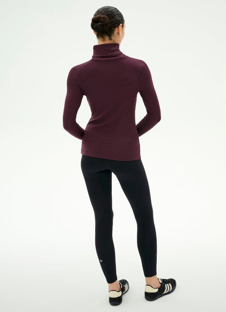 A woman shown from the back wearing a fitted long-sleeve turtleneck top in deep burgundy, paired with black leggings and black-and-white athletic shoes, against a plain white studio background.