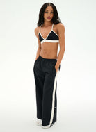 A model stands against a plain white studio background wearing a black and white sports bra and matching black wide-leg track pants with white side stripes.