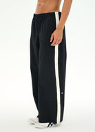 A person wearing loose black track pants with a bold white vertical stripe down the side, paired with white sneakers, shown against a plain white studio background.