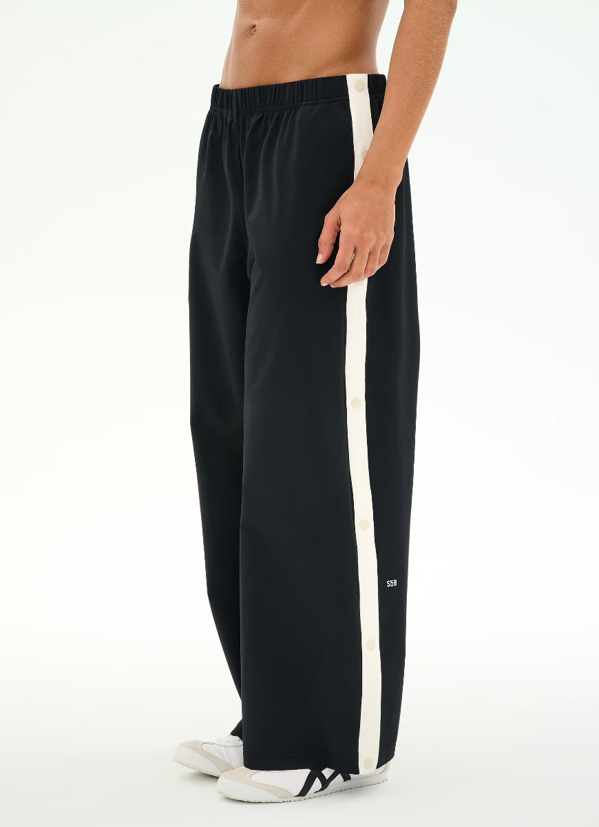 A person wearing loose black track pants with a bold white vertical stripe down the side, paired with white sneakers, shown against a plain white studio background.