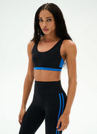 A woman modeling a black athletic set consisting of a sports bra and high-waisted leggings with bright blue side accents, photographed against a clean white studio background.