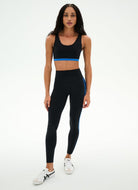 A woman modeling a matching black activewear set consisting of a sports bra with a bright blue band and high-waisted leggings with blue side stripes, paired with white sneakers, against a plain white studio background.