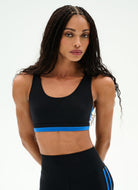 A woman modeling a black athletic sports bra with a bright blue band detail across the underbust, paired with matching black athletic leggings featuring blue side stripes, against a clean white background.