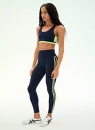 A woman modeling a matching navy blue activewear set consisting of a sports bra and high-waisted leggings, both featuring lime green side accents, paired with white sneakers.