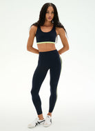 A woman modeling a matching athletic set consisting of a dark navy sports bra with a neon yellow-green trim and high-waisted leggings with a matching side stripe, paired with white sneakers, against a plain white studio background.