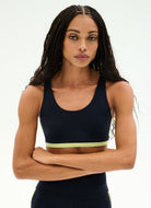 A woman modeling a dark navy athletic sports bra with a light yellow-green underband, posed against a clean white studio background.