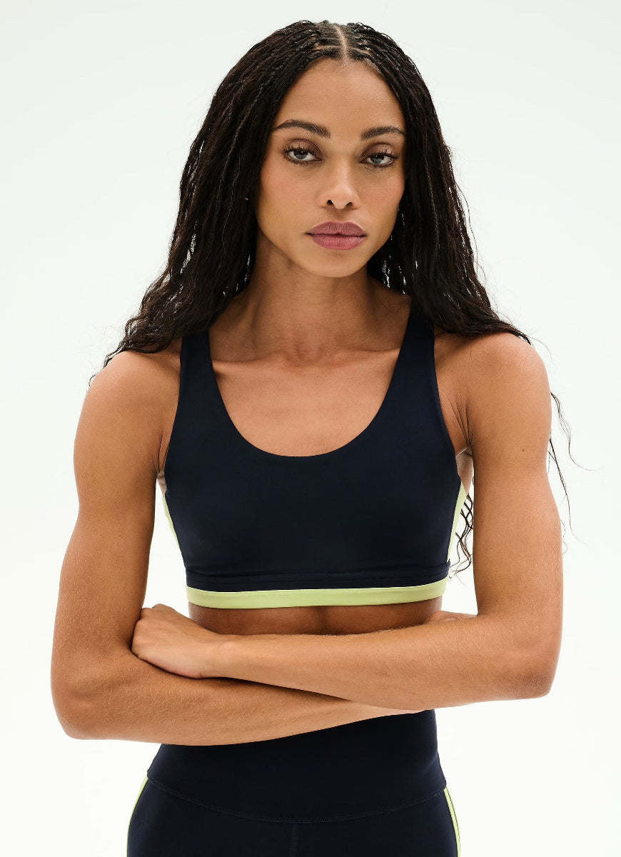 A woman modeling a dark navy athletic sports bra with a light yellow-green underband, posed against a clean white studio background.