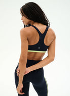 A woman shown from the back wearing a dark navy athletic sports bra with a light yellow-green band and matching dark leggings with a thin light yellow-green stripe, against a clean white studio background.