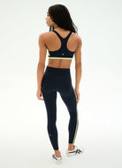 A woman shown from the back wearing a dark navy athletic set consisting of a sports bra with a neon yellow-green band and matching high-waisted leggings with a neon yellow-green side stripe, paired with white sneakers.