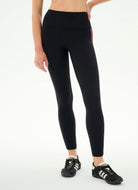 A model wearing black high-waisted leggings with a thin red stripe down the side, paired with black sneakers, shown from the waist down against a plain white background.