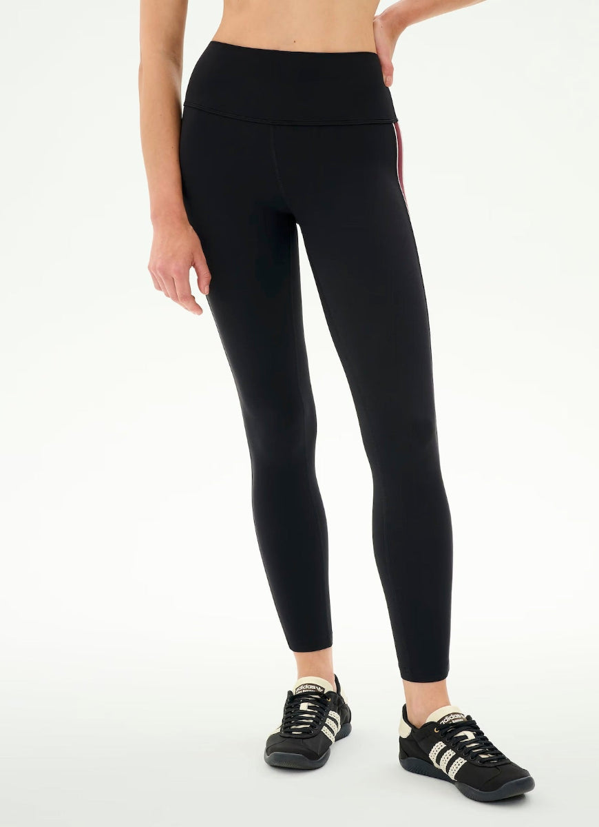 A model wearing black high-waisted leggings with a thin red stripe down the side, paired with black sneakers, shown from the waist down against a plain white background.