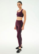 A woman modeling a matching athletic set consisting of a scoop-neck sports bra and high-waisted leggings in deep plum purple with thin black side stripes, paired with black sneakers, against a plain white studio background.