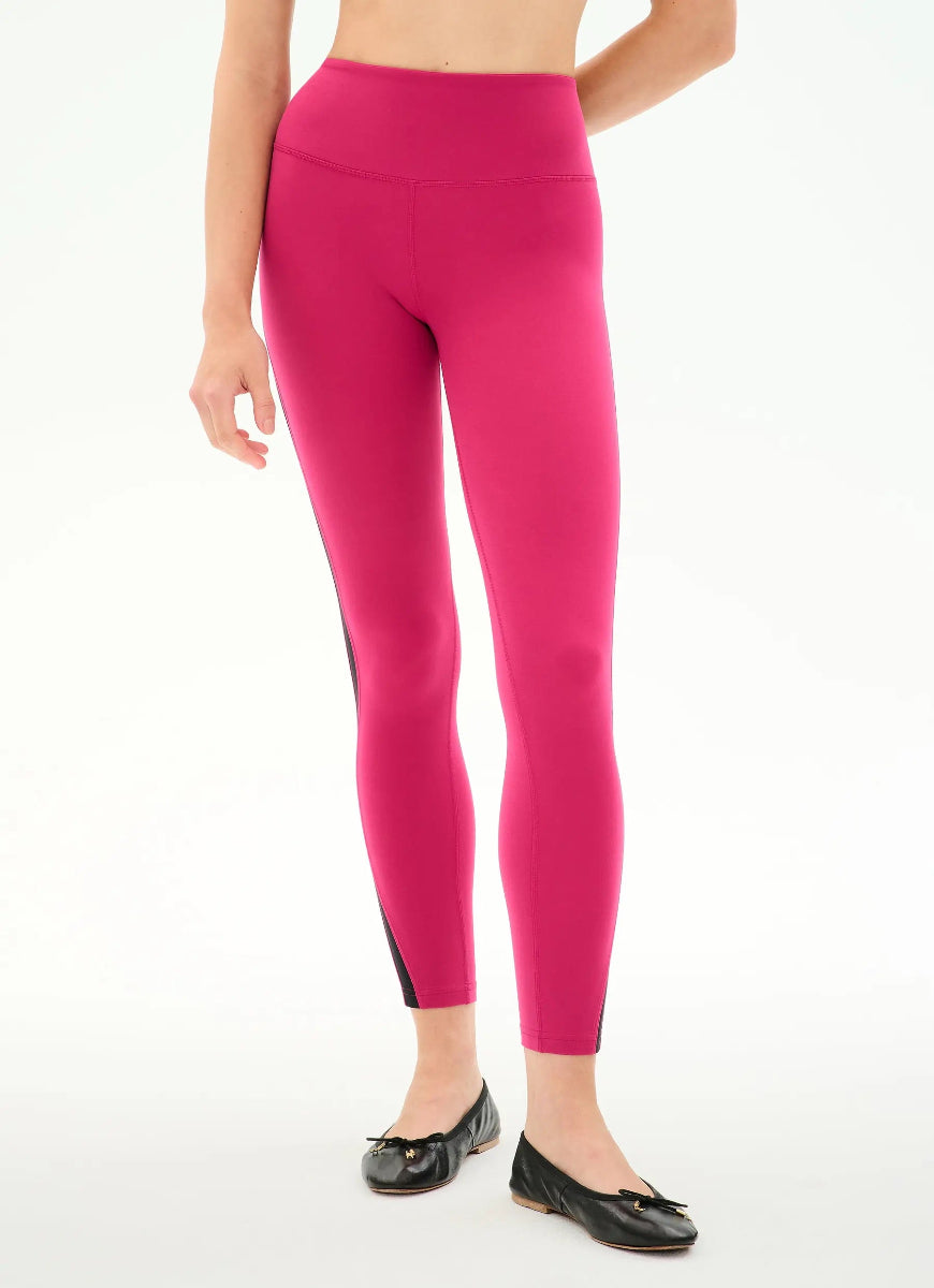 A model wearing bright pink high-waisted leggings with black side panels and black flat shoes, shown from the waist down on a white studio background.